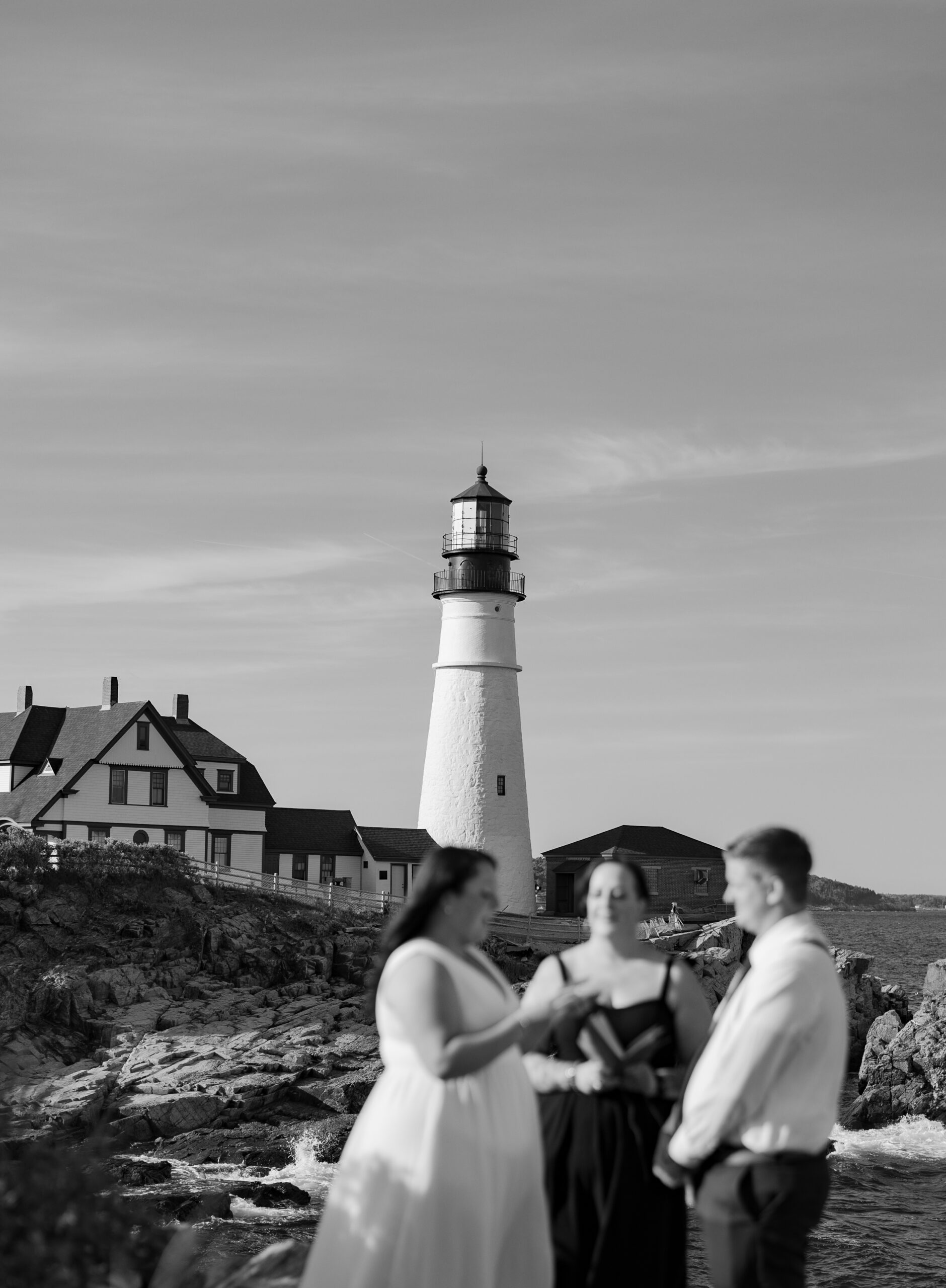 Black and white photo of a Maine elopement at Portland Head Light with bride, groom, and officiant blurred in the foreground and the lighthouse in sharp focus in the background