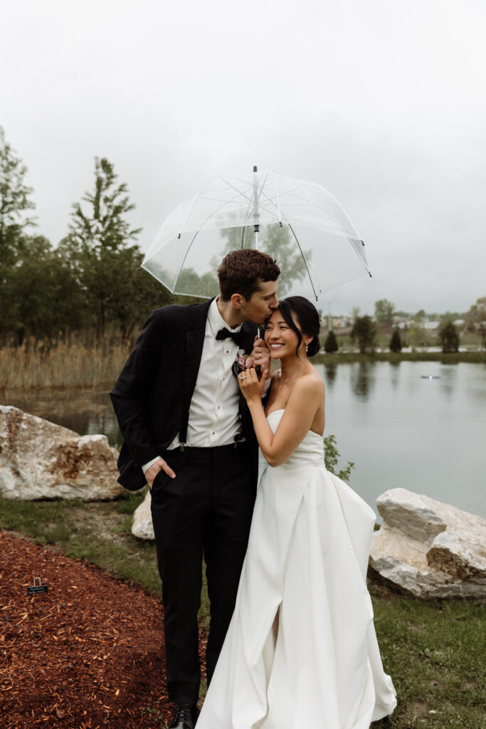 Bride and groom embracing beneath a transparent umbrella as gentle rain falls at the Essex Resort and Spa, Vermont wedding venue.
