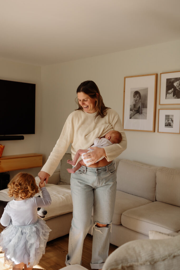 Maine family photographed in their cozy living room during an in-home session. Mother wearing cream colors twirls her daughter and holds her newborn.