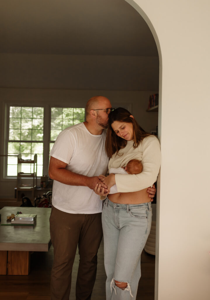 Maine family in-home session: mother holds newborn while father kisses her on the head in their farmhouse kitchen