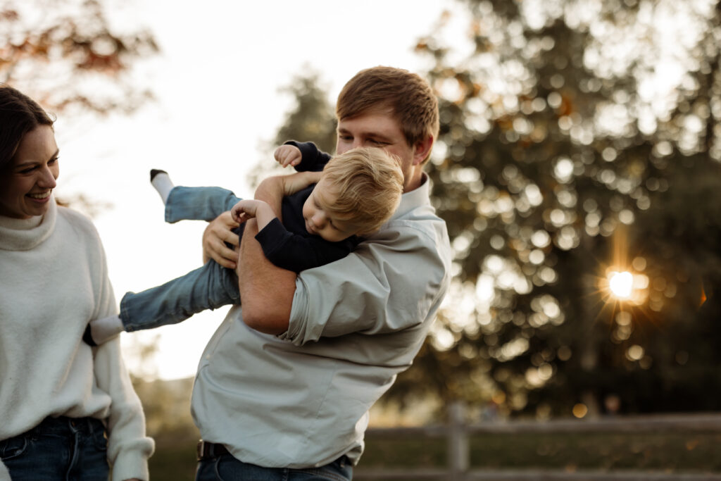 Father holding his son playfully in the air while laughing during family photoshoot in the summer. The mother smiles at the son.