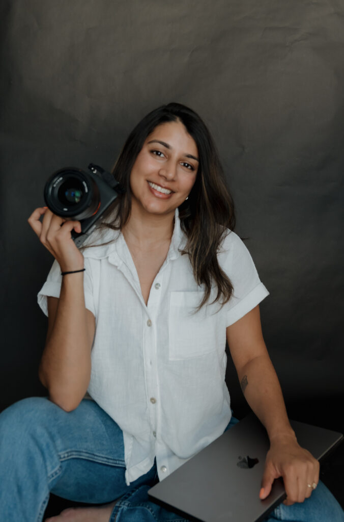 Headshot of Maine family photographer Damini Fulmer sitting against a black backdrop, smiling while holding a camera in one hand and a laptop in the other, wearing a white short-sleeve linen button-down and jeans, hair down