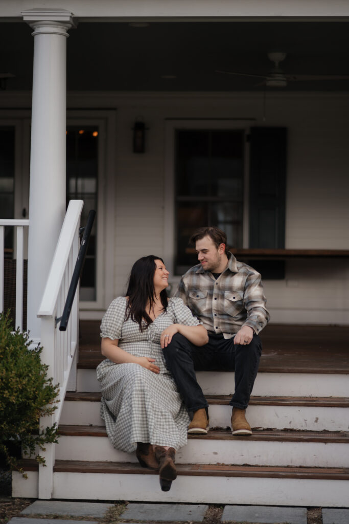 Maine maternity photographer captures expecting couple smiling at each other on front porch steps, pregnant wife at 36 weeks in a long flowing dress