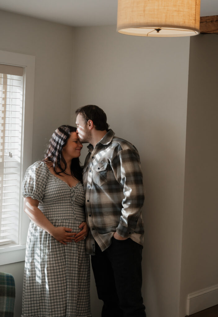 Expecting father kissing pregnant wife’s forehead in intimate indoor maternity portrait